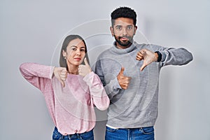 Young hispanic couple standing together doing thumbs up and down, disagreement and agreement expression