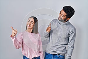 Young hispanic couple standing together looking proud, smiling doing thumbs up gesture to the side