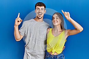 Young hispanic couple standing together over blue background smiling amazed and surprised and pointing up with fingers and raised