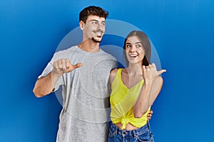 Young hispanic couple standing together over blue background smiling with happy face looking and pointing to the side with thumb