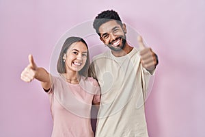 Young hispanic couple together over pink background approving doing positive gesture with hand, thumbs up smiling and happy for