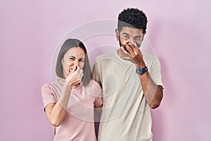 Young hispanic couple together over pink background smelling something stinky and disgusting, intolerable smell, holding breath