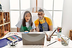 Young hispanic couple using laptop sitting on the table at art studio scared and amazed with open mouth for surprise, disbelief