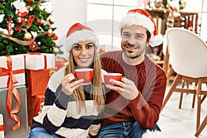 Young hispanic couple wearing christmas hat drinking coffee at home