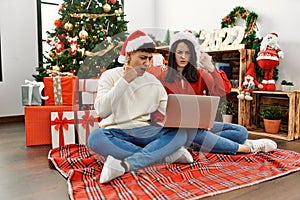Young hispanic couple wearing christmas hat using laptop sitting by christmas tree annoyed and frustrated shouting with anger,