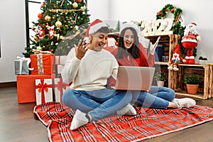 Young hispanic couple wearing christmas hat using laptop sitting by christmas tree celebrating victory with happy smile and winner