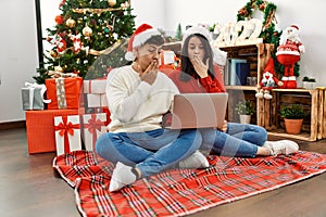 Young hispanic couple wearing christmas hat using laptop sitting by christmas tree covering mouth with hand, shocked and afraid