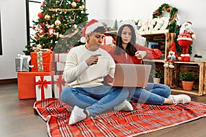 Young hispanic couple wearing christmas hat using laptop sitting by christmas tree pointing with finger to the camera and to you,