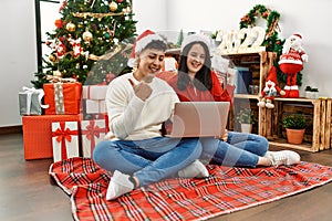Young hispanic couple wearing christmas hat using laptop sitting by christmas tree pointing thumb up to the side smiling happy