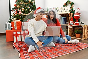 Young hispanic couple wearing christmas hat using laptop sitting by christmas tree scared and amazed with open mouth for surprise,