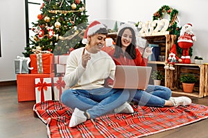 Young hispanic couple wearing christmas hat using laptop sitting by christmas tree smiling happy pointing with hand and finger to