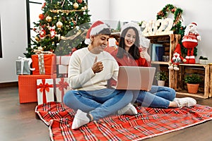 Young hispanic couple wearing christmas hat using laptop sitting by christmas tree smiling happy and positive, thumb up doing