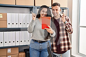 Young hispanic couple working at small business ecommerce doing video call smiling happy pointing with hand and finger to the side