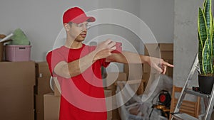 Young hispanic courier with smartphone points finger inside building interior amid stacks of moving boxes; focus