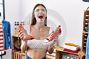 Young hispanic customer woman holding high heel shoes and sneaker angry and mad screaming frustrated and furious, shouting with