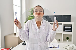 Young hispanic dentist woman holding ordinary toothbrush and electric toothbrush puffing cheeks with funny face