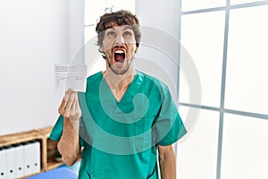 Young hispanic doctor man holding covid record card angry and mad screaming frustrated and furious, shouting with anger