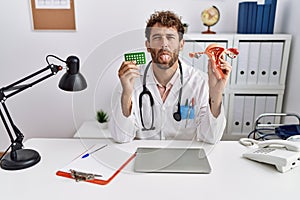 Young hispanic doctor man holding female genital organ and birth control pills sticking tongue out happy with funny expression