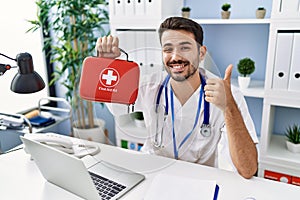 Young hispanic doctor man holding first aid kit smiling happy and positive, thumb up doing excellent and approval sign