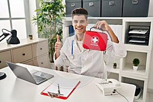 Young hispanic doctor man holding first aid kit smiling happy and positive, thumb up doing excellent and approval sign