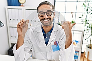 Young hispanic doctor man holding medical hearing aid pointing thumb up to the side smiling happy with open mouth