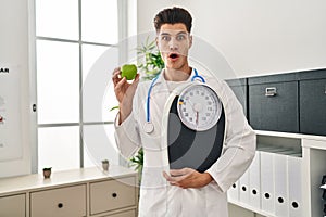 Young hispanic doctor man holding scale at dietitian clinic afraid and shocked with surprise and amazed expression, fear and