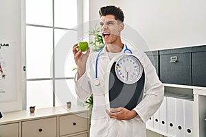 Young hispanic doctor man holding scale at dietitian clinic angry and mad screaming frustrated and furious, shouting with anger