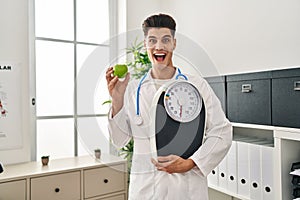 Young hispanic doctor man holding scale at dietitian clinic celebrating crazy and amazed for success with open eyes screaming