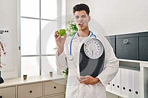 Young hispanic doctor man holding scale at dietitian clinic clueless and confused expression