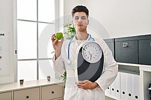 Young hispanic doctor man holding scale at dietitian clinic looking at the camera blowing a kiss being lovely and sexy