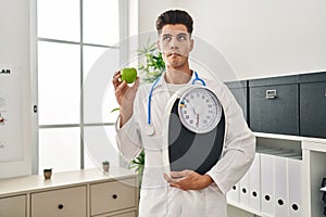 Young hispanic doctor man holding scale at dietitian clinic making fish face with mouth and squinting eyes, crazy and comical