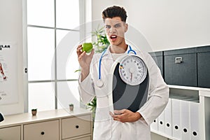 Young hispanic doctor man holding scale at dietitian clinic in shock face, looking skeptical and sarcastic, surprised with open