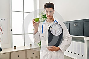 Young hispanic doctor man holding scale at dietitian clinic skeptic and nervous, frowning upset because of problem