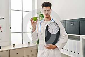 Young hispanic doctor man holding scale at dietitian clinic skeptic and nervous, frowning upset because of problem