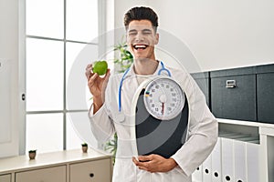 Young hispanic doctor man holding scale at dietitian clinic smiling and laughing hard out loud because funny crazy joke