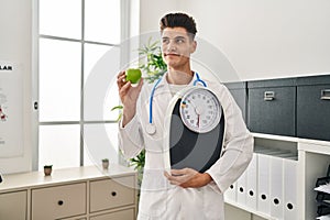 Young hispanic doctor man holding scale at dietitian clinic smiling looking to the side and staring away thinking