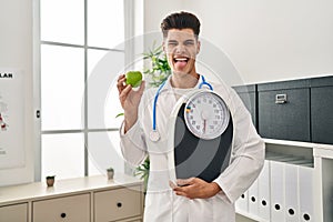 Young hispanic doctor man holding scale at dietitian clinic sticking tongue out happy with funny expression