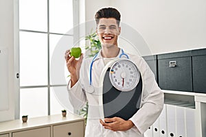 Young hispanic doctor man holding scale at dietitian clinic winking looking at the camera with sexy expression, cheerful and happy