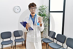 Young hispanic doctor man at waiting room pointing to the back behind with hand and thumbs up, smiling confident