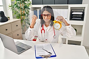 Young hispanic doctor woman holding alarm clock at the clinic annoyed and frustrated shouting with anger, yelling crazy with anger