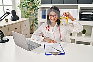 Young hispanic doctor woman holding alarm clock at the clinic looking positive and happy standing and smiling with a confident