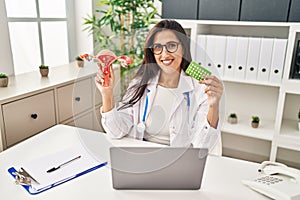 Young hispanic doctor woman holding anatomical female genital organ and birth control pills smiling with a happy and cool smile on