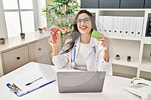 Young hispanic doctor woman holding anatomical female genital organ and birth control pills smiling and laughing hard out loud
