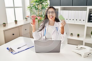 Young hispanic doctor woman holding anatomical female genital organ and birth control pills sticking tongue out happy with funny