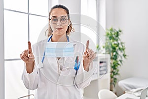 Young hispanic doctor woman holding safety mask puffing cheeks with funny face