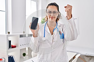 Young hispanic doctor woman holding smartphone showing screen annoyed and frustrated shouting with anger, yelling crazy with anger