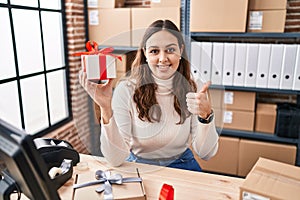 Young hispanic doctor woman working at small business ecommerce holding present smiling happy and positive, thumb up doing