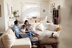 Young Hispanic family sitting on sofa reading a book together in the living room, seen from doorway