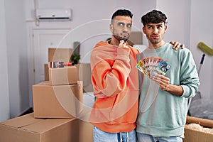 Young hispanic gay couple moving to a new home holding banknotes serious face thinking about question with hand on chin,