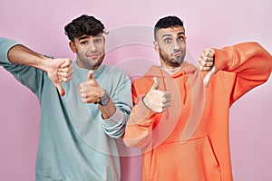 Young hispanic gay couple standing over pink background doing thumbs up and down, disagreement and agreement expression
